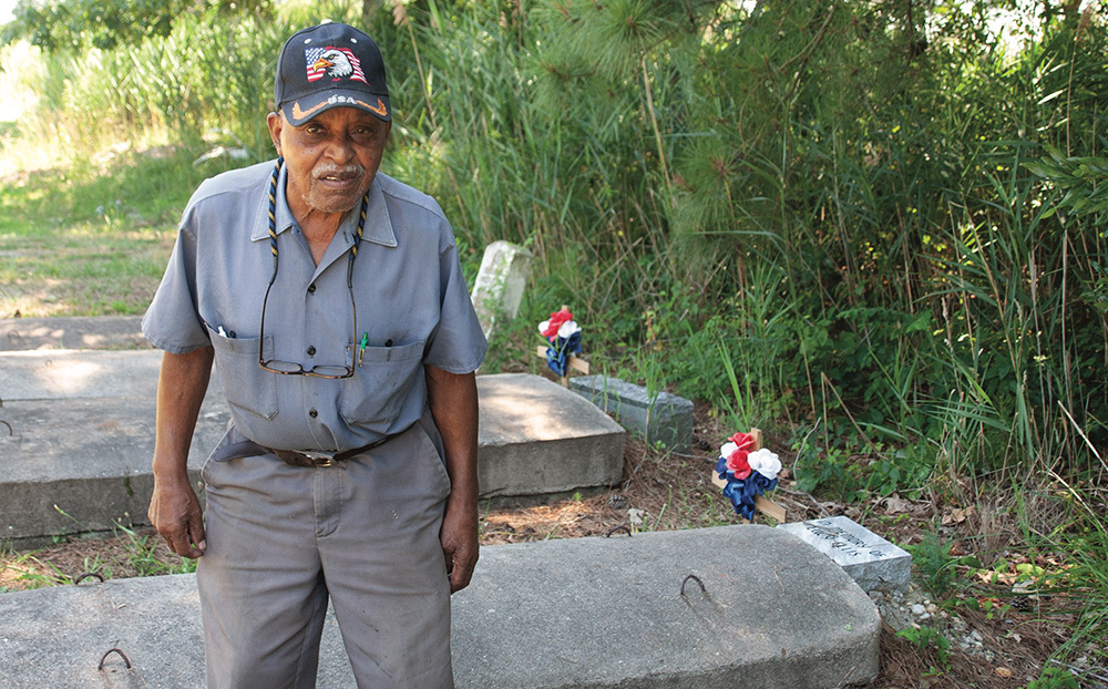 Luther Cornish stands in the cemetery across from his home. Beyond the tall marsh grass is water. Photo, Nicole Lehming / MDSG