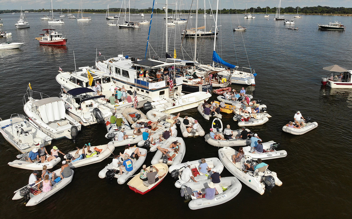 Aerial image of approximately 30 boats on the water, tied together. Approximately 40 boats are sailing close by. There are trees in the distance. Photo, ShoreRivers